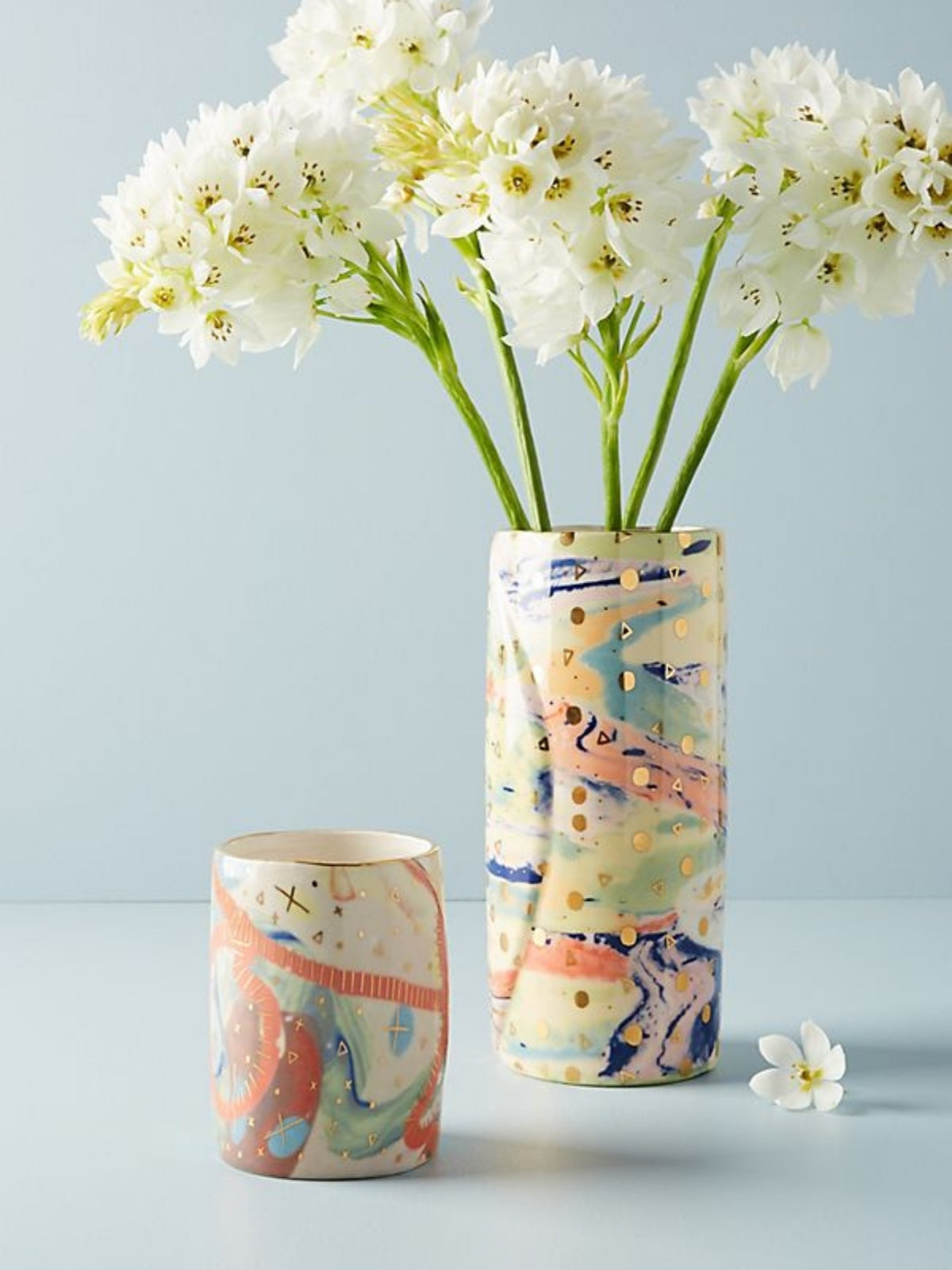 Two ceramic vases with abstract multicolor patterns and gold leaf accents, one tall vase holding white flowers and one smaller cup, set against a light blue background.