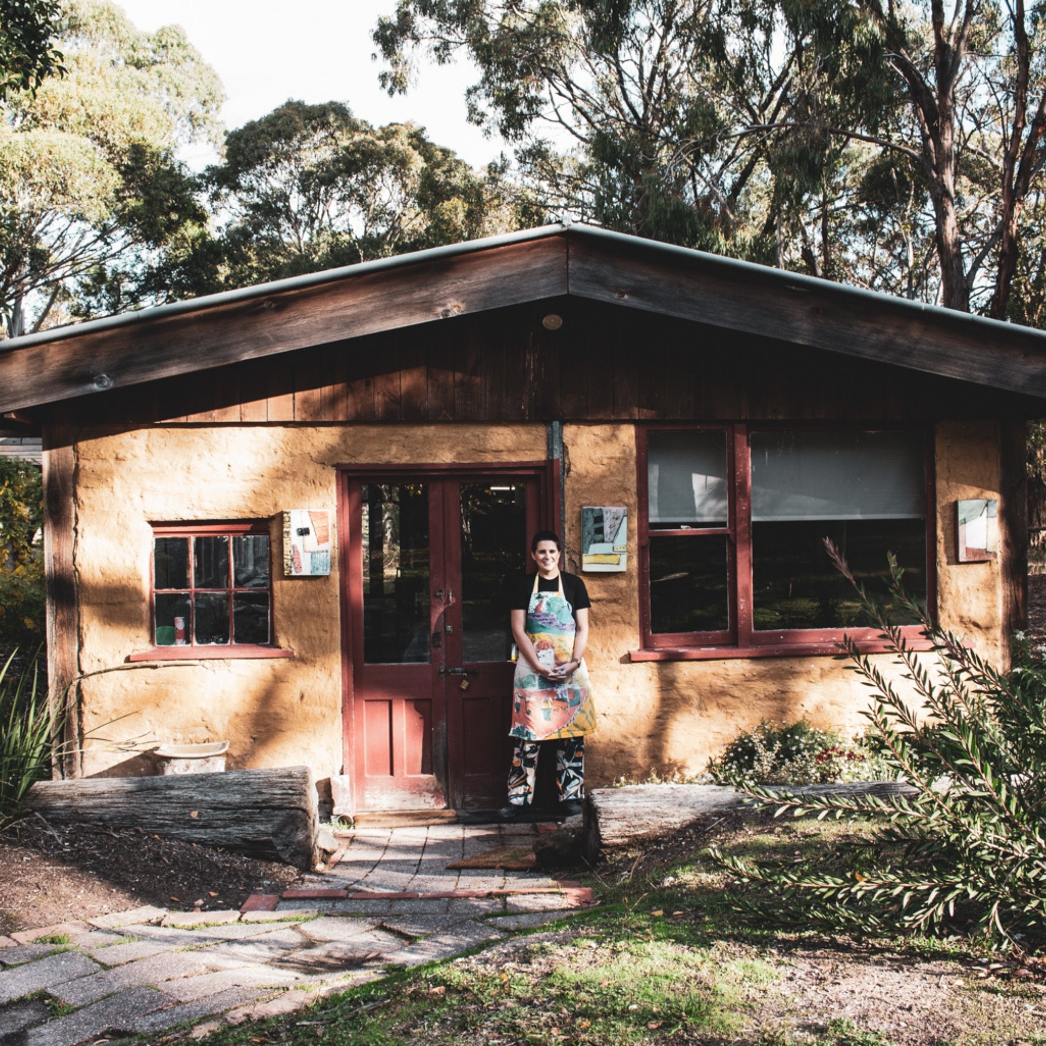 A person wearing a colorful apron stands in front of a rustic wooden and stucco cabin surrounded by trees.